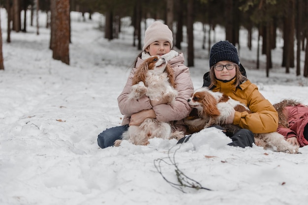 Três meninas adoráveis se divertindo juntas pela bela floresta congelada do parque Irmãs fofas brincando na neve com um cachorro