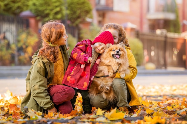 Três meninas duas irmãs mais velhas e uma criança estão andando com um pomeranian fofo em um dia ensolarado de outono a garotinha abraça e acaricia o cachorro