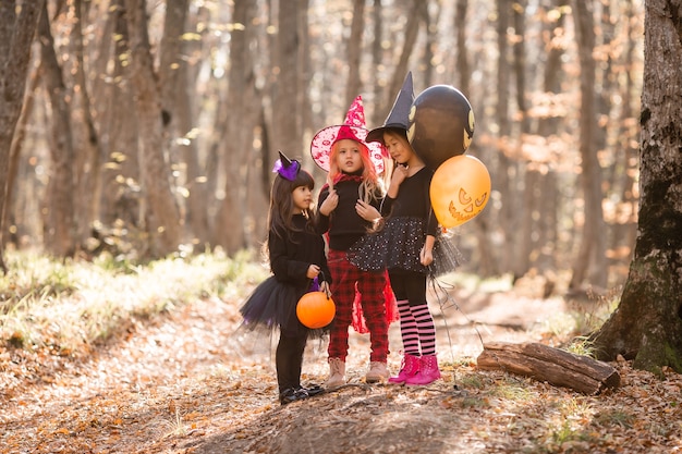 Três meninas em fantasias de bruxas riem caminhando pela floresta de outono com cestas de doces