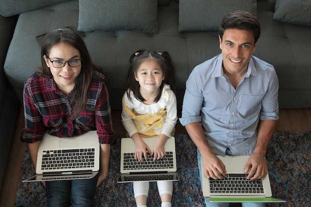 Tres miembros de una familia diversa, padre caucásico y madre asiática y media hija pequeña sentados juntos en la sala de estar de la casa y usando 3 computadoras portátiles. Idea para trabajar en casa.