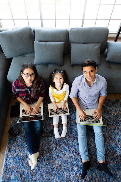 Tres miembros de una familia diversa, padre caucásico y madre asiática y media hija pequeña sentados juntos en la sala de estar de la casa y usando 3 computadoras portátiles. Idea para trabajar en casa.