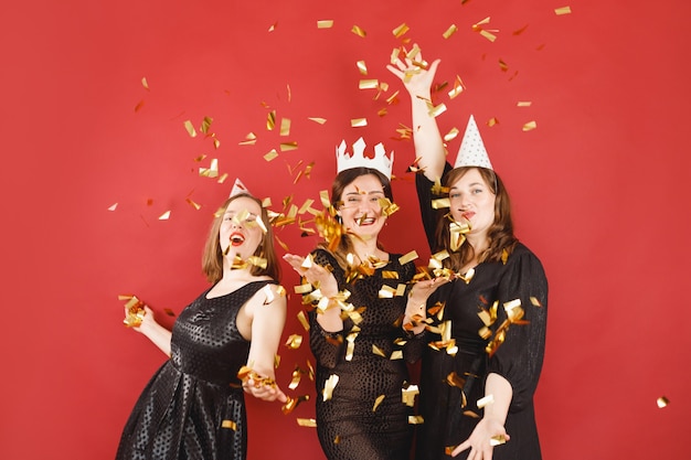 Tres mujeres encantadas en vestido negro y sombreros de fiesta sobre fondo rojo con confeti dorado. Fiesta femenina