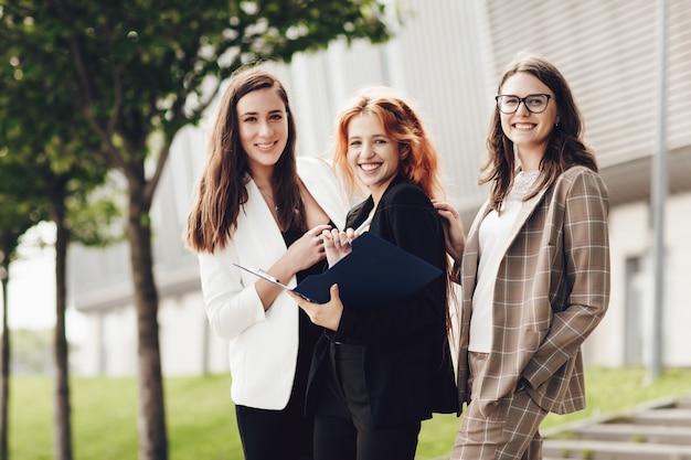 Tres mujeres jóvenes con estilo que trabajan al aire libre y sonriendo, sostienen una carpeta con documentos
