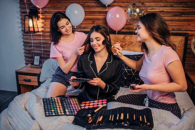 Tres mujeres jóvenes felices se sientan en la cama en la sala festiva. Dos chicas hacen cuidado del cabello y maquillaje para la tercera. Hay cepillos y sombra en la cama. Las mujeres en pijama negro sostienen un conjunto de sombras en las manos.