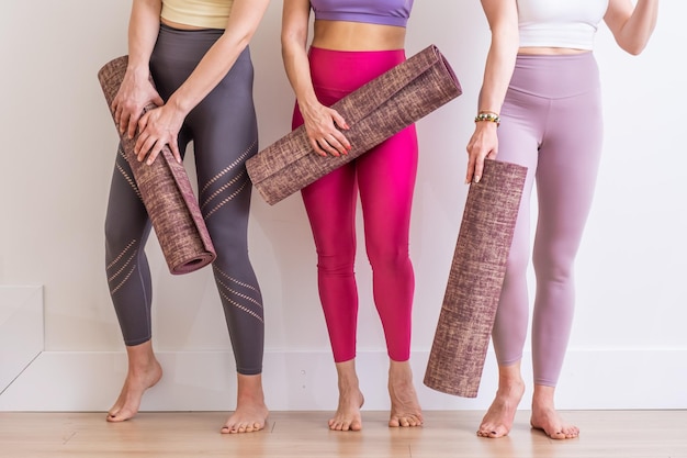 Tres mujeres jóvenes sosteniendo colchoneta de yoga en ropa deportiva colorida en entrenamiento de profesor de yoga de estudio blanco