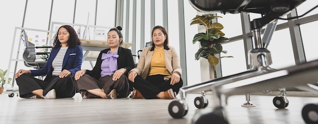 Tres mujeres de negocios asiáticas sentadas en el suelo en la oficina con varias ventanas de cristal meditando junto a las mesas de conferencias.