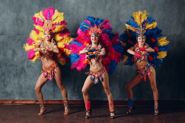 Três mulher dançando em traje de carnaval de samba brasileiro com plumagem de penas coloridas.