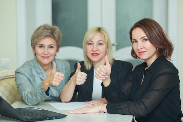 Três mulheres adultas de terno estão sentadas à mesa com um laptop no trabalho no escritório com um gesto de mãos