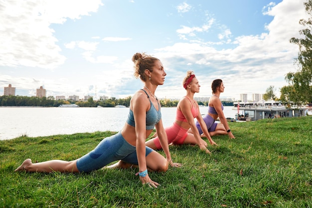 Três mulheres bonitas em forma fazendo Virabhadrasana malhando no parque no dia de verão vestindo tops de roupas esportivas conceito de estilo de vida de comprimento total