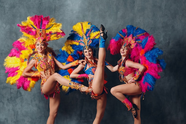 Três mulheres em traje de cabaré com plumagem de penas coloridas.