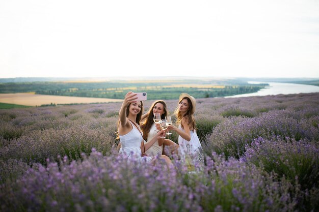 Três mulheres em vestidos brancos bebem champanhe em um campo de lavanda e tiram selfie