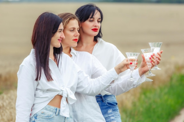 Três mulheres felizes desfrutando de taças de vinho branco e sorrindo durante o piquenique Melhores amigas meninas bonitas e elegantes bebem vinho e riem Amigos de comemoração de aniversário brindam com champanhe