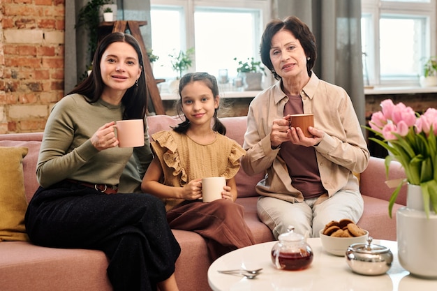 Três mulheres felizes tomando chá enquanto estão sentadas no sofá ao lado da mesa servida