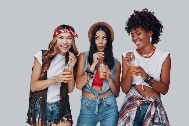 Três mulheres jovens e atraentes bebendo um coquetel refrescante e sorrindo em pé contra um fundo cinza