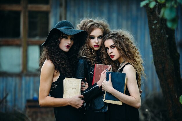 Três mulheres vintage como bruxas posam em frente a um prédio abandonado com livros nas mãos na véspera do Halloween