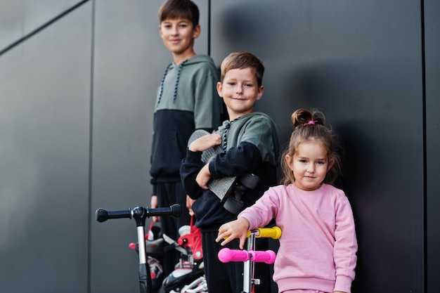 Tres niños al aire libre contra la pared moderna negra La familia deportiva pasa tiempo libre al aire libre con scooters y patines