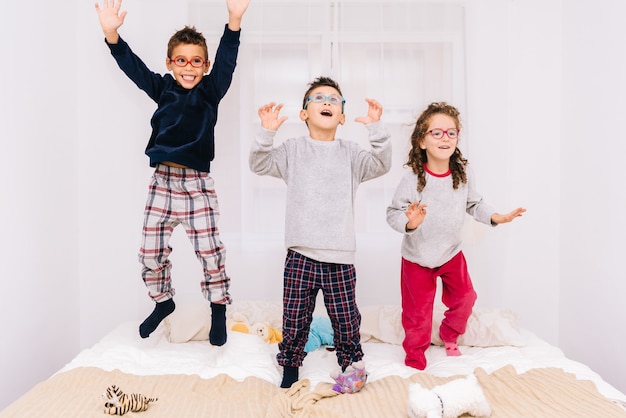 Tres niños alegres con gafas saltando y jugando en la cama