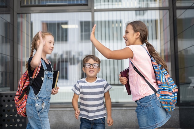 Tres niños caucásicos lindos felices con anteojos y mochila se paran y hablan juntos, dan cinco en el patio de la escuela. Vacaciones escolares. Nerd. Día del conocimiento. De cerca. Concepto de regreso a la escuela.