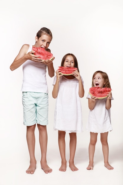 Tres niños en el estudio se paran sobre un fondo blanco y comen trozos de sandía Hermano dos hermanas con ropas brillantes descalzas Mimos de la infancia feliz Fuente de alimentación adecuada