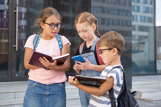 Tres niños y una mochila se paran y leen un libro, con un teléfono en el patio de la escuela. De vuelta a la escuela