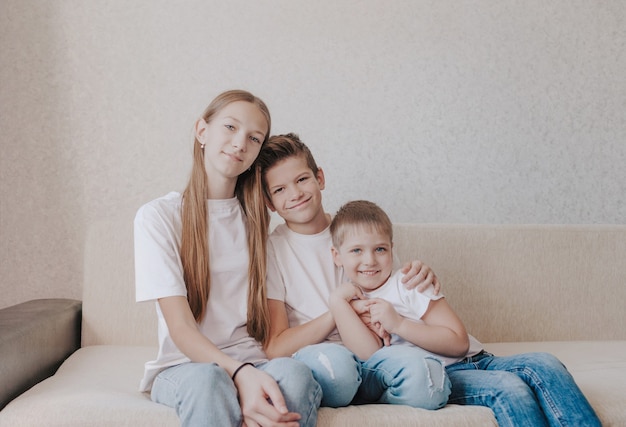 Tres niños, una niña y dos niños con camisetas blancas y jeans azules, se sientan abrazados en el sofá de la casa y sonríen.