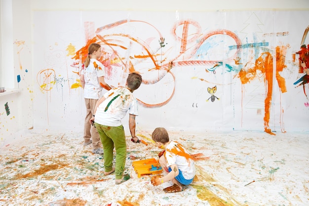 Tres niños pintan en una pared blanca con pinturas de niños y niñas.