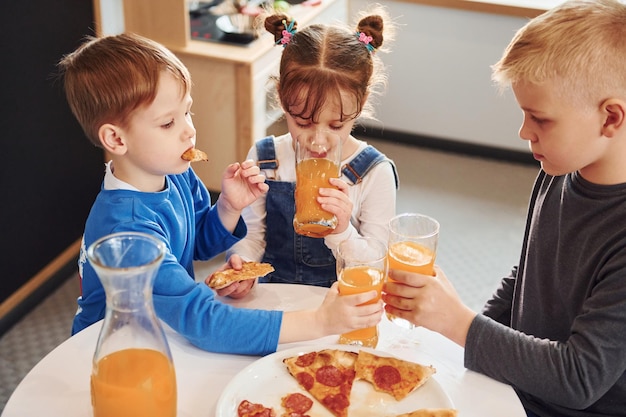 Tres niños sentados en el interior junto a la mesa y comiendo pizza con jugo de naranja juntos