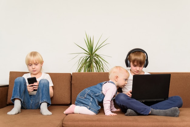 Tres niños en el sofá con teléfono y ordenador portátil. Alpha Generation y tecnología moderna. Estilo de vida moderno con dispositivo electrónico.