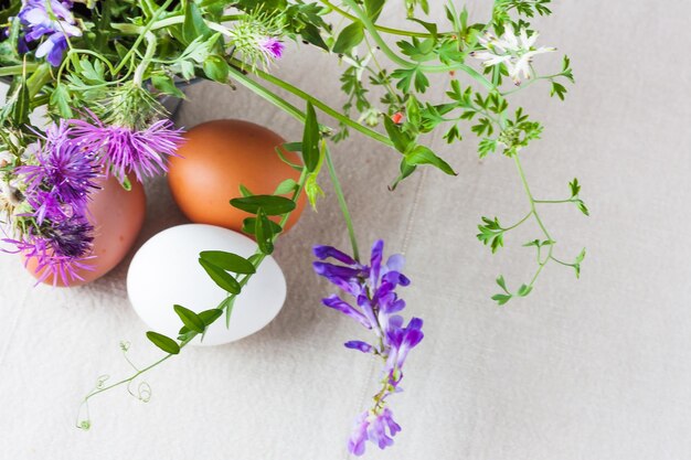 Foto três ovos de galinha multicoloridos em uma toalha de mesa de linho com um buquê de flores silvestres de primavera