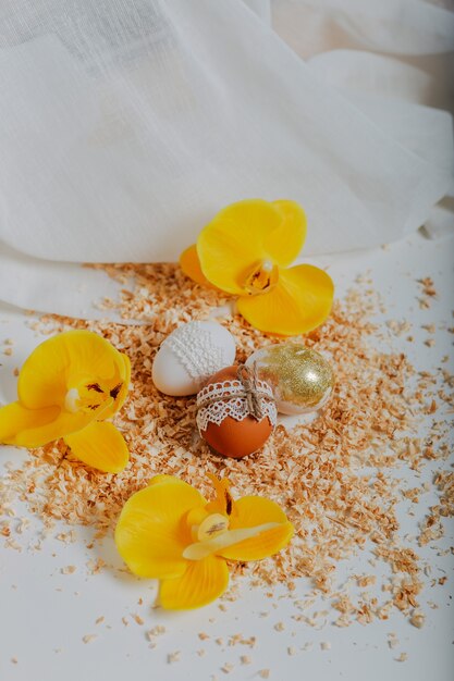 Três ovos de páscoa em fundo branco com serragem ao redor. cartão de páscoa feliz com flores amarelas da orquídea. decoração de ovos de páscoa com borda de renda e corda. ovo de páscoa dourado.