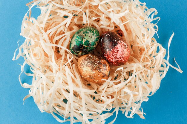 Três ovos de páscoa são pintados em vermelho, amarelo e verde. Os ovos estão em um ninho de aparas de madeira. Ovos de Páscoa pintados sobre um fundo azul. Postura plana. Fechar-se.