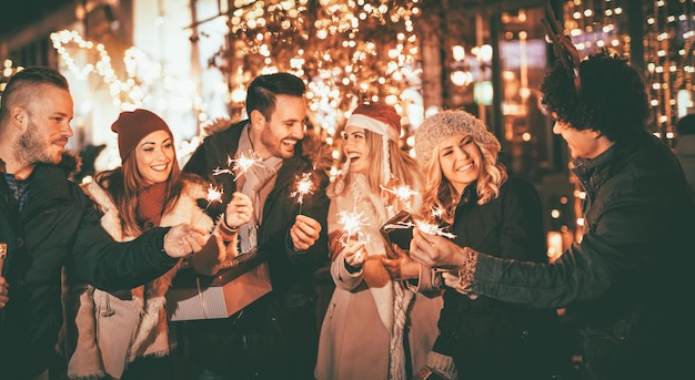 Tres parejas con bengalas disfrutando de la fiesta de Navidad al aire libre en la calle de la ciudad por la noche y con muchas luces de fondo.