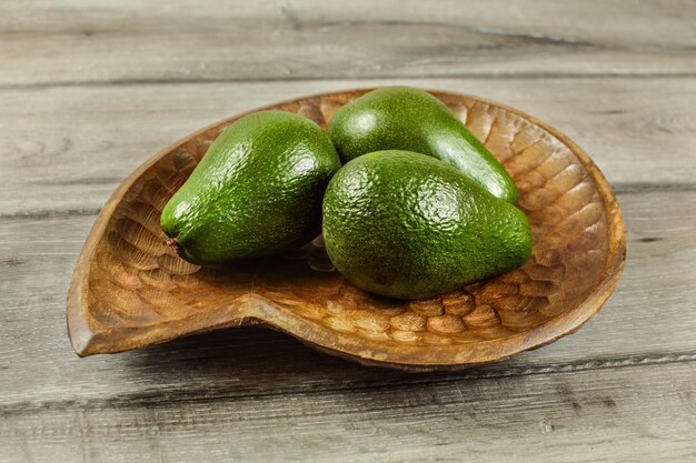 Tres peras de aguacate enteras en un tazón de madera, sobre una mesa rústica.