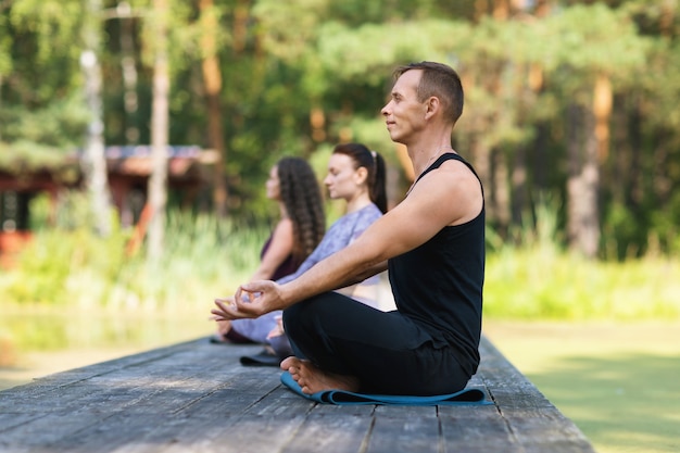 Três pessoas estão meditando sentadas em uma esteira na posição de lótus no parque