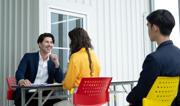 Três pessoas estão sentadas em uma mesa com um homem de terno e gravata sorrindo para os outros dois