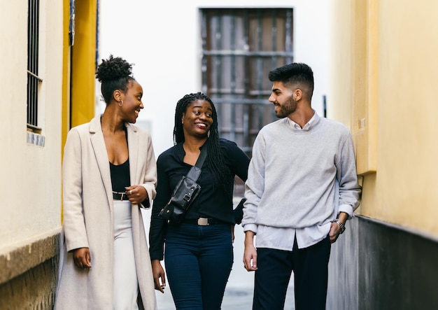 Três pessoas multiétnicas com roupas da moda conversando animadamente em uma rua estreita