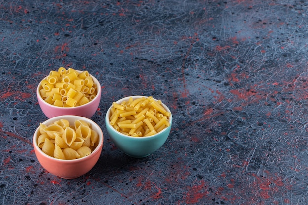 Tres platos de colores con diferentes tipos de pasta cruda sobre un fondo oscuro.