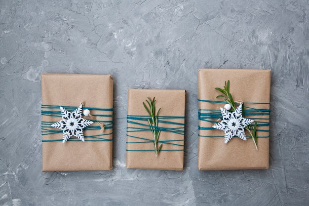 Três presentes de Natal em papel ofício embrulhado em fio azul, com flocos de neve brancos e ramos de alecrim, sobre um fundo cinza de concreto.