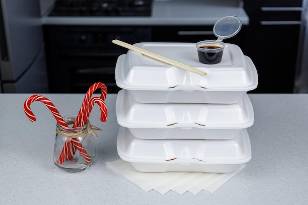 Três recipientes de comida branca estão na mesa da cozinha Pauzinhos para comer sushi e uma caixa de molho de soja Doces vermelhos de Natal em uma jarra