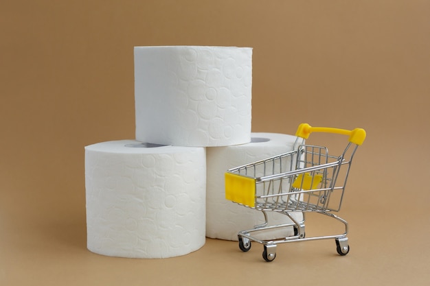 Tres rollos de papel higiénico blanco sobre fondo blanco y un pequeño carrito de supermercado