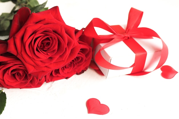 Três rosas vermelhas e uma caixa de presente com um laço em uma mesa branca e corações vermelhos.