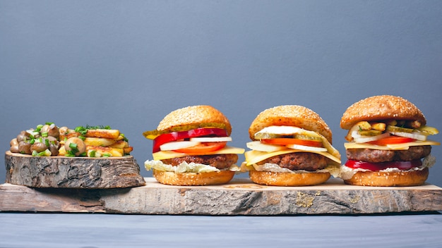 Tres sabrosas hamburguesas variadas con papas fritas y champiñones espolvoreados con cebollas verdes en los posavasos de madera del bosque sobre un fondo gris. Almuerzo abundante.