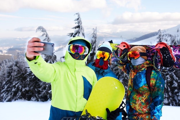 Três snowboarders tirando selfie com câmera de smartphone na estância de esqui amigos fotografando para rede social compartilhando com pranchas de snowboard perto da floresta usando óculos reflexivos roupas de moda coloridas