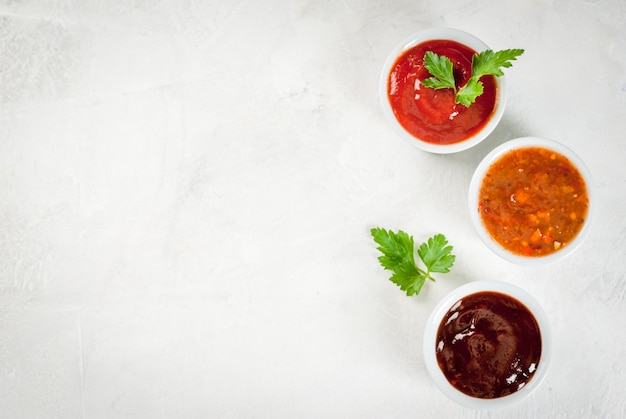 Três tipos de molho de tomate vermelho em uma mesa de pedra branca: ketchup clássico tradicional, molho barbecue, molho chinês agridoce. Espaço de cópia da vista superior