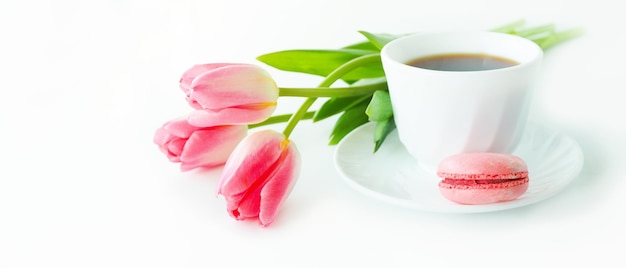 Três tulipas cor de rosa, macarons e uma xícara de café em uma mesa branca.