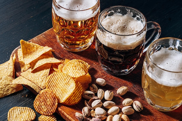 Tres vasos de cerveza artesanal cara, clásica y sin filtrar y oscura en un vaso sobre la mesa con un refrigerio de chips de maní y pistacho y nachos