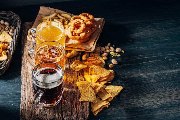 Tres vasos de cerveza artesanal cara, clásica y sin filtrar y oscura en un vaso sobre la mesa con un refrigerio de chips de maní y pistacho y nachos