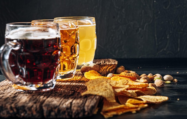 Tres vasos de cerveza artesanal cara, clásica y sin filtrar y oscura en un vaso sobre la mesa con un refrigerio de chips de maní y pistacho y nachos