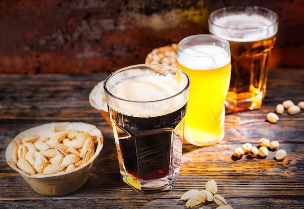 Tres vasos con cerveza clara, sin filtrar y oscura se colocan en una fila cerca de platos con bocadillos y nueces esparcidas en un escritorio de madera oscura. Concepto de alimentos y bebidas