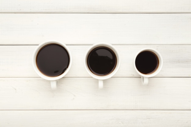 Três xícaras de café de tamanhos diferentes com café expresso preto forte. preparando bebidas em fundo branco de madeira, vista superior. conceito de energia e refresco, copie o espaço